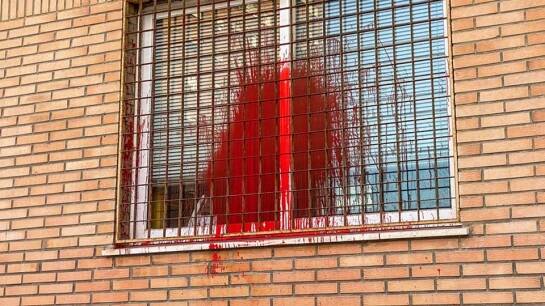 A picture of a window that someone has thrown red paint over. The window is barred with black iron rods and a brown brick wall surrounds it 